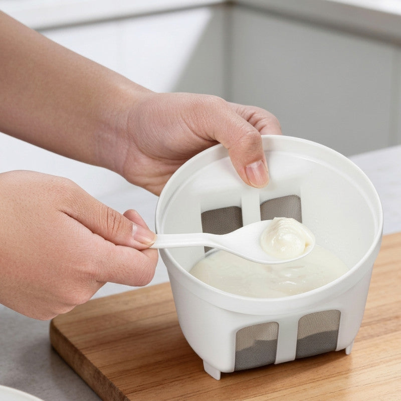 A close-up of a person's hands using a white strainer bowl with mesh filters to separate a creamy white liquid, set on a wooden cutting board.